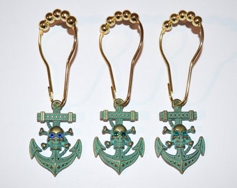 May include: Three shower curtain hooks with a gold finish and teal anchor pendants. Each anchor pendant features a skull and crossbones design with jewel-toned eyes. The hooks have a decorative gold bead detail at the top.
