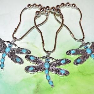 May include: Set of three silver-tone shower curtain hooks with dragonfly pendants. Each dragonfly features turquoise-colored stones and intricate detailing. The hooks have a decorative beaded top.