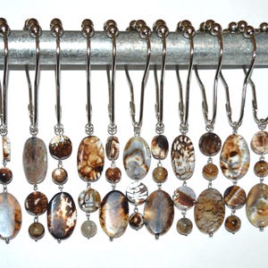 May include: Shower curtain hooks with decorative oval and round beads in shades of brown, tan, and cream. The beads are strung on silver-colored metal hooks, designed to hang from a shower rod. The hooks are a decorative bathroom accessory.