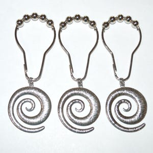 May include: Three silver-toned shower curtain hooks, each featuring a metallic spiral pendant. The hooks are designed with a rounded top section adorned with five small silver spheres, providing a functional mechanism for hanging curtains.