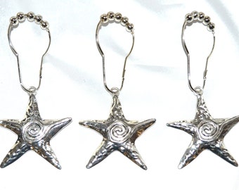 May include: Three silver-colored shower curtain hooks. Each hook features a starfish pendant with a textured surface and a spiral design in the center. The hooks have a series of small, round beads at the top.