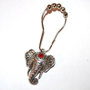 May include: Silver-toned shower curtain hook with an elephant head pendant. The elephant pendant has detailed carvings and a red jewel in the center. The hook has a series of small silver beads at the top.
