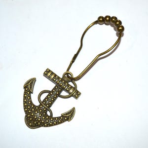 May include: A bronze-toned metal shower curtain ring featuring a decorative anchor charm. The anchor has a textured, dotted surface and is attached to a wire loop that includes four small decorative beads at the top for sliding along a rod.