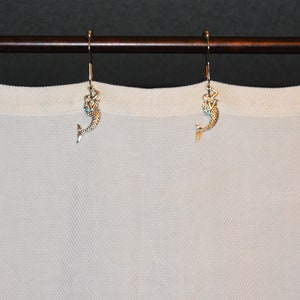 Puede incluir: Un conjunto de ganchos decorativos para cortinas con forma de sirena. Los ganchos son de color plateado con figuras detalladas de sirenas, colgando de una barra marr&oacute;n. Se ve una cortina blanca en el fondo.