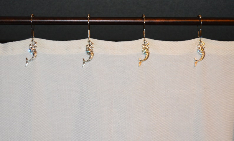 Puede incluir: Un conjunto de ganchos decorativos para cortinas con forma de sirena. Los ganchos son de color plateado con figuras detalladas de sirenas, colgando de una barra marr&oacute;n. Se ve una cortina blanca en el fondo.