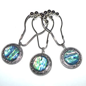May include: Three silver-toned shower curtain hooks, each featuring a circular pendant with an iridescent abalone shell inlay. The metal hooks have a smooth, curved design with small decorative beads at the top for sliding along a rod.