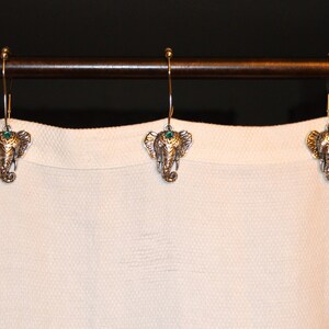 May include: Three decorative shower curtain hooks with an elephant head design. The hooks are gold-colored with a detailed elephant head and a small green gem. They hang from a brown metal rod against a white curtain.