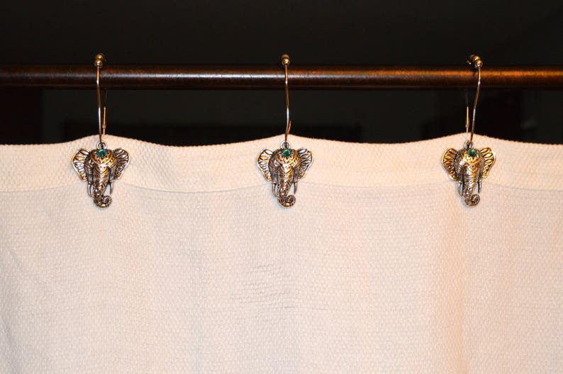 May include: Three decorative shower curtain hooks with an elephant head design. The hooks are gold-colored with a detailed elephant head and a small green gem. They hang from a brown metal rod against a white curtain.