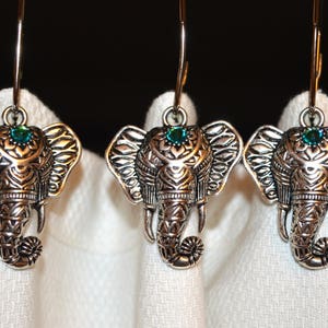 May include: Three silver-toned elephant earrings with intricate detailing. Each elephant has a small teal-colored gem on its forehead. The earrings are suspended from thin, silver-colored hooks, and are displayed against a white background.