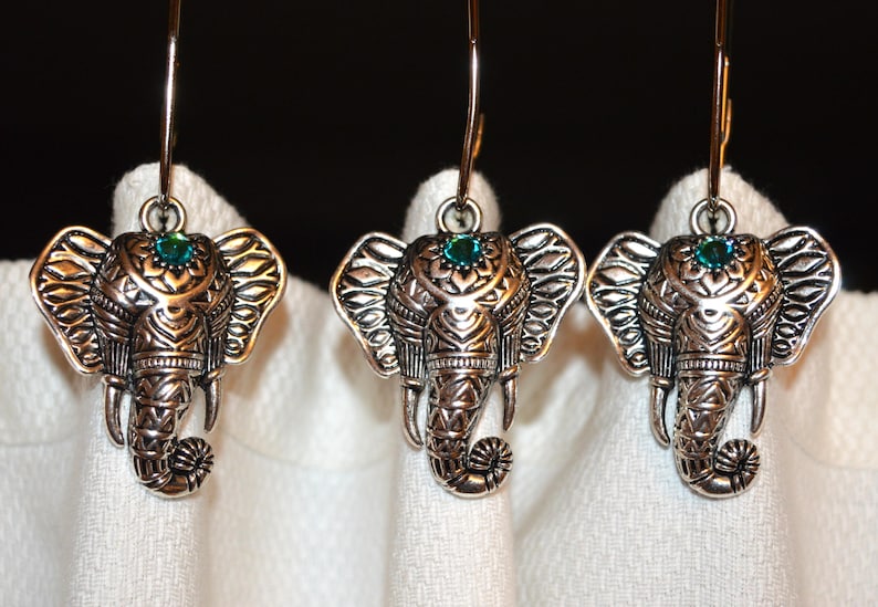 May include: Three silver-toned elephant earrings with intricate detailing. Each elephant has a small teal-colored gem on its forehead. The earrings are suspended from thin, silver-colored hooks, and are displayed against a white background.