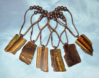May include: A set of six shower curtain hooks featuring polished copper-toned metal rings with spherical beads and dangling, rectangular-shaped tiger's eye gemstone pendants. Each natural stone displays unique brown and gold banded patterns.