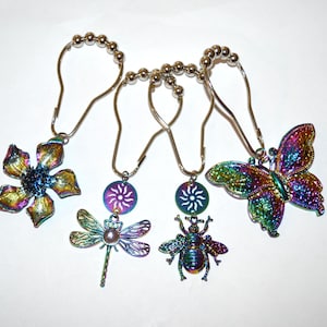 May include: Set of five iridescent shower curtain hooks. Each hook features a different charm: a flower, a dragonfly, a sunburst, a bee, and a butterfly. The charms have a metallic finish with colors of blue, green, and gold.
