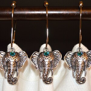 May include: Three silver-colored elephant-shaped shower curtain hooks. Each hook features an elephant head with detailed carvings and a teal-colored gem. The hooks hang from a dark brown rod against a white curtain.
