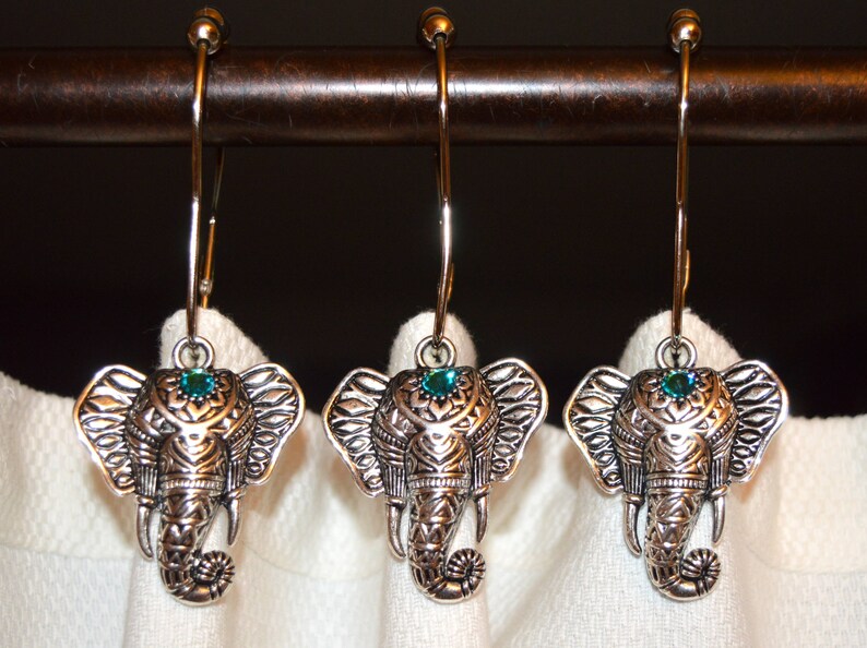 May include: Three silver-colored elephant-shaped shower curtain hooks. Each hook features an elephant head with detailed carvings and a teal-colored gem. The hooks hang from a dark brown rod against a white curtain.