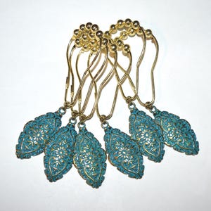 May include: Set of six decorative shower curtain hooks. Each hook features a gold-colored metal design with a teal-colored, ornate, leaf-shaped pendant. The pendants have a detailed floral pattern and are suspended from gold-colored hooks.