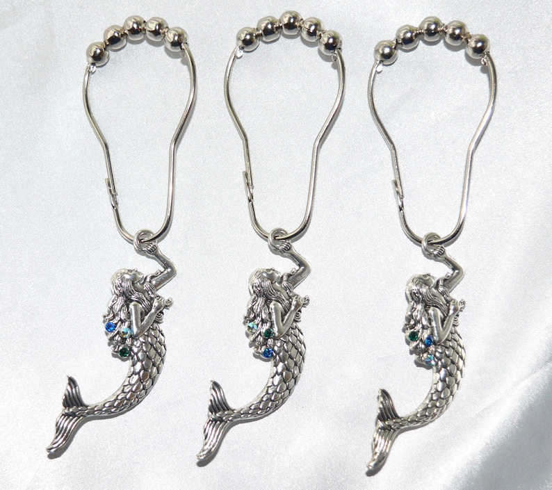 Puede incluir: Tres ganchos plateados para cortinas de ducha con dijes de sirena. Cada gancho tiene una serie de peque&ntilde;as cuentas plateadas en la parte superior y un colgante de sirena con detalles azules y verdes. Las sirenas tienen cabello suelto y escamas detalladas.