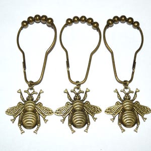 May include: Three antique brass-toned shower curtain hooks, each featuring a detailed bee charm attached to a metal loop with five small decorative spheres at the top. The hooks have a teardrop-shaped frame and a hinged closure mechanism.