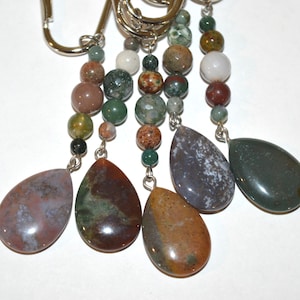 May include: Close-up of a keychain with multiple strands of beads and teardrop-shaped pendants. The beads are various sizes and colors, including green, brown, and white. The pendants are polished stones in shades of brown, green, and purple.