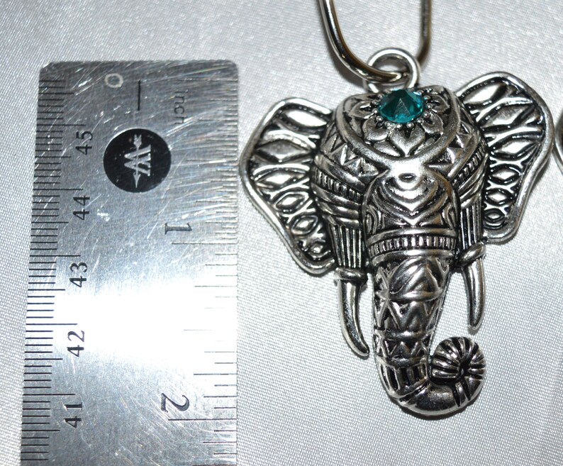 May include: Silver-toned elephant pendant with intricate detailing, a teal-colored gem, and a loop for hanging. The pendant is approximately 1.75 inches in length, showcasing a detailed design. The elephant's trunk is curled, and the ears are adorned with patterns.