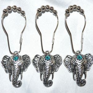 May include: Three silver-colored shower curtain hooks with elephant pendants. Each elephant has detailed carvings and a turquoise-colored gem. The hooks have a beaded top and a curved design for easy hanging. The background is a white, silky fabric.