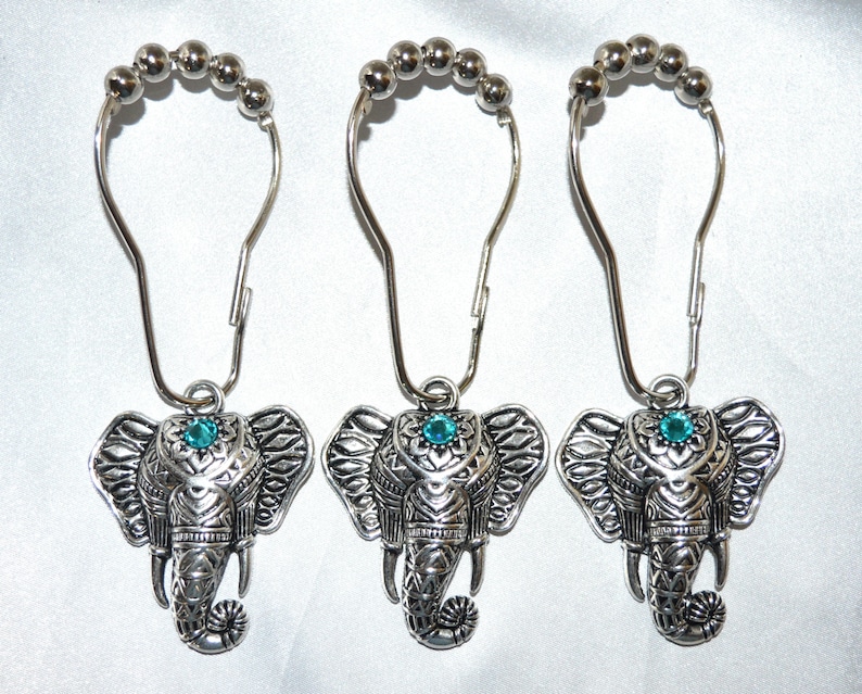 May include: Three silver-colored shower curtain hooks with elephant pendants. Each elephant has detailed carvings and a turquoise-colored gem. The hooks have a beaded top and a curved design for easy hanging. The background is a white, silky fabric.