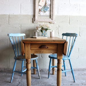 May include: A wooden drop-leaf table with a single central drawer featuring a dark metal pull, accompanied by two light blue spindle-back chairs. The table has four turned legs and a natural wood finish, while the chairs show signs of wear.