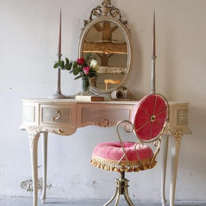 May include: A vintage dressing table set featuring an oval mirror, a pink and gold vanity table, and a matching pink velvet chair. The chair has a gold frame and tassel trim. Two tall candles and a vase of flowers are on the table.
