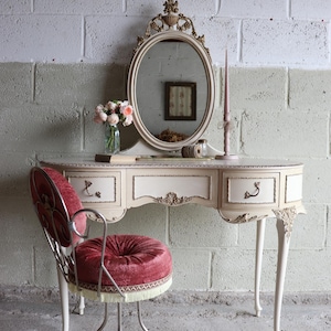 May include: A cream-coloured vanity table with an oval mirror, featuring ornate gold-toned carvings and three drawers. A matching chair with a circular, tufted, deep red velvet seat and a metal frame with fringe trim sits in front of the table.