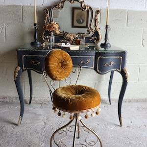 May include: A vanity chair with a mustard yellow tufted velvet seat and backrest, featuring a gold metal frame with scrollwork and a decorative pom-pom fringe trim, positioned in front of a black vanity table with gold accents and a mirror.
