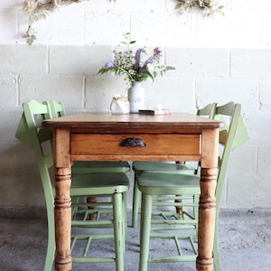 May include: A rectangular wooden dining table with a natural finish and a single central drawer featuring a dark metal pull. The table is supported by four turned legs and is accompanied by four light green wooden chairs with slatted backs.