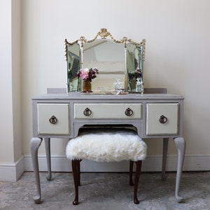 May include: A grey vanity table with three drawers and a white cushioned stool. The table has a tri-fold mirror with a gold frame and ornate details. The mirror is positioned above the table.