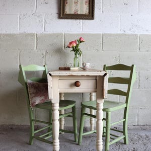 May include: A vintage-style cream table with a drawer and two green chairs. A vase of pink roses and a framed floral print are on the wall. The table is approximately 76 cm high.