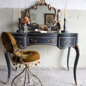 May include: A black vanity desk with gold trim, three drawers, and a matching mirror with an ornate gold frame. A swivel chair with a round, tufted olive green velvet seat and backrest, featuring a decorative fringe trim and metal legs.