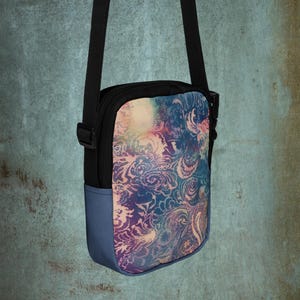 May include: A small, black crossbody bag with a colorful paisley pattern in shades of blue, purple, and pink. The bag has a blue bottom and a black adjustable strap. The bag is designed for carrying essentials.