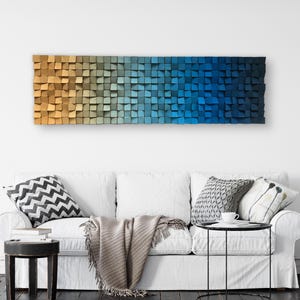 May include: A long, rectangular wall art piece made of wood blocks in shades of blue, green, and brown. The blocks are arranged in a gradient pattern, starting with a light brown color and transitioning to a deep blue color. The art piece is hanging on a white wall above a white couch with pillows and a throw blanket.