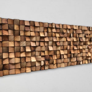 Può includere: Un'opera d'arte da parete in legno con un effetto 3D. Il legno è di colore marrone chiaro e i pezzi sono disposti a forma di onda.