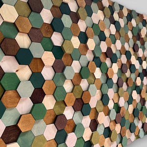 Könnte beinhalten: Ein Wandkunstwerk aus zahlreichen sechseckigen Holzfliesen. Die Fliesen sind in einem Mosaikmuster angeordnet und weisen eine Vielzahl von Farben auf, darunter Grüntöne, Brauntöne und natürliche Holztöne. Das Kunstwerk erzeugt einen strukturierten, dreidimensionalen Effekt.