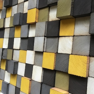 Può includere: Un'opera d'arte murale realizzata con blocchi di legno in varie tonalità di grigio, bianco e giallo. I blocchi sono disposti a scalare, creando una texture visivamente interessante.