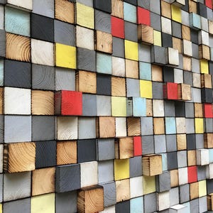 Puede incluir: Una obra de arte mural compuesta por numerosos bloques de madera en varios colores, incluyendo rojo, amarillo, gris, negro y tonos de madera natural. Los bloques están dispuestos en un patrón de cuadrícula, creando un efecto texturizado y tridimensional.