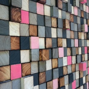 May include: A wall art piece composed of various wooden blocks in shades of gray, brown, pink, and white. The blocks are arranged in a grid pattern, creating a textured, three-dimensional effect. The wood grain is visible on many of the blocks.