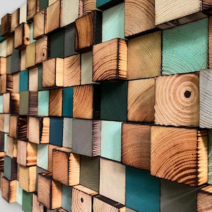 Puede incluir: Una pieza de arte mural compuesta por numerosos bloques de madera de diversas profundidades y tamaños. Los bloques presentan vetas de madera natural, bordes carbonizados y superficies pintadas en tonos verde azulado, menta, verde oscuro, gris y marrón claro.