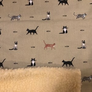 May include: A beige fabric with a repeating pattern of black, white, and gray cats. The fabric has a brown and white sherpa lining.