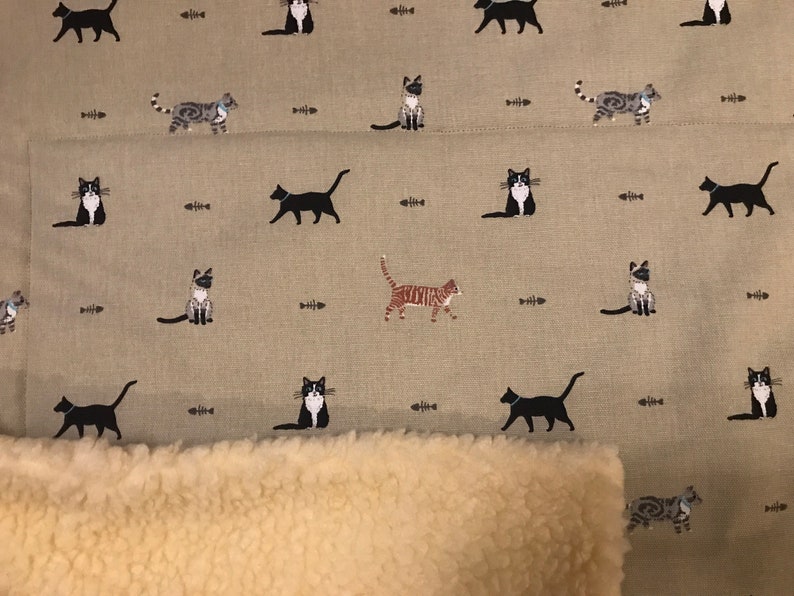May include: A beige fabric with a repeating pattern of black, white, and gray cats. The fabric has a brown and white sherpa lining.