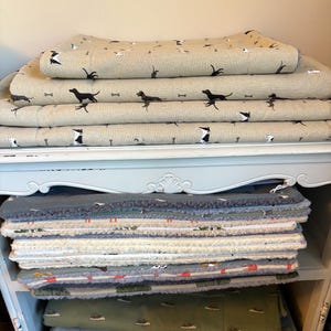 May include: A stack of dog beds and blankets. The top layers feature a tan fabric with black dog silhouettes and bone patterns. Below, a variety of patterned and solid-colored beds are visible, including green and gray options. The items are stacked on a light blue shelf.