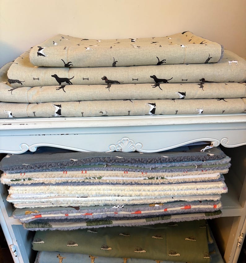 May include: A stack of dog beds and blankets. The top layers feature a tan fabric with black dog silhouettes and bone patterns. Below, a variety of patterned and solid-colored beds are visible, including green and gray options. The items are stacked on a light blue shelf.