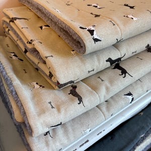 May include: A stack of beige pet beds with a repeating pattern of black and white dog illustrations and bone shapes. The beds have a soft, gray, plush lining. The beds are folded and stacked on top of each other.