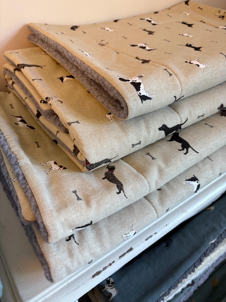 May include: A stack of beige pet beds with a repeating pattern of black and white dog illustrations and bone shapes. The beds have a soft, gray, plush lining. The beds are folded and stacked on top of each other.