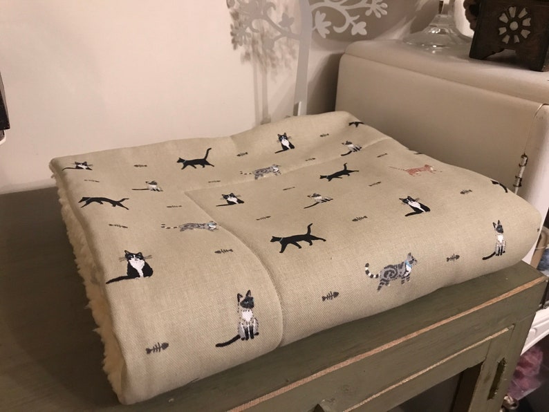 May include: A beige throw blanket with a pattern of black, white, and gray cats. The blanket is folded and sitting on a wooden table.