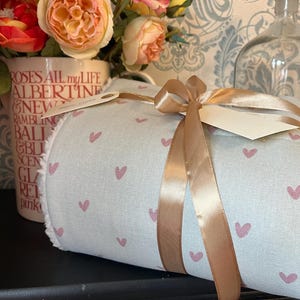 Puede incluir: Una manta enrollada con un fondo azul claro y un patrón de corazones rosas, atada con una cinta dorada. Se adjunta una etiqueta en blanco. La manta está sobre una superficie negra, con flores y objetos decorativos en el fondo.