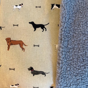 May include: A beige fabric with a repeating pattern of various dog breeds and bone shapes. The fabric is next to a fluffy, light blue material. The dogs are in black, brown, and white. The overall theme is pet-related.